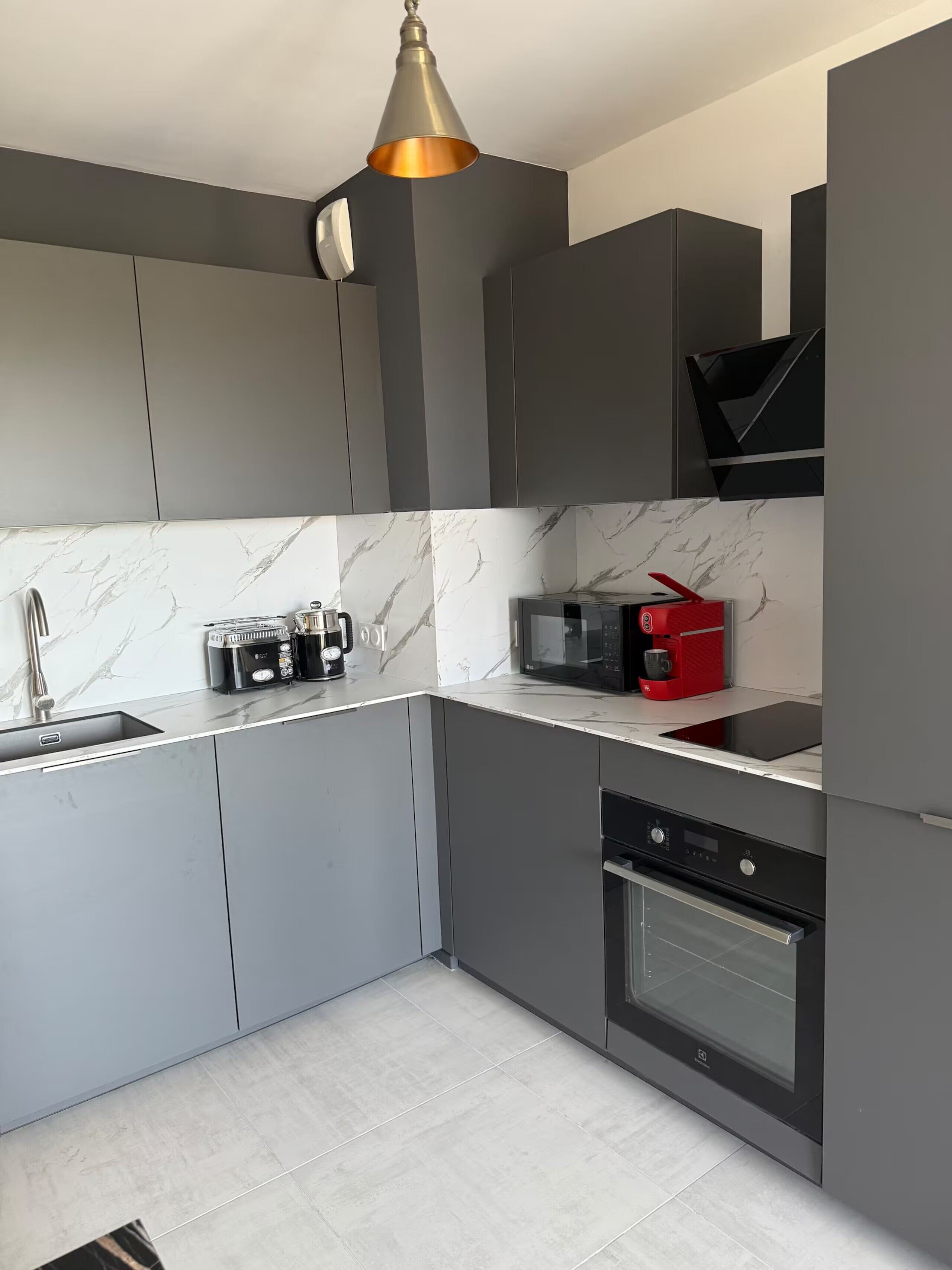 A modern kitchen with grey cabinets, a marble backsplash, a built-in microwave, a red coffee machine, and a stainless steel oven.