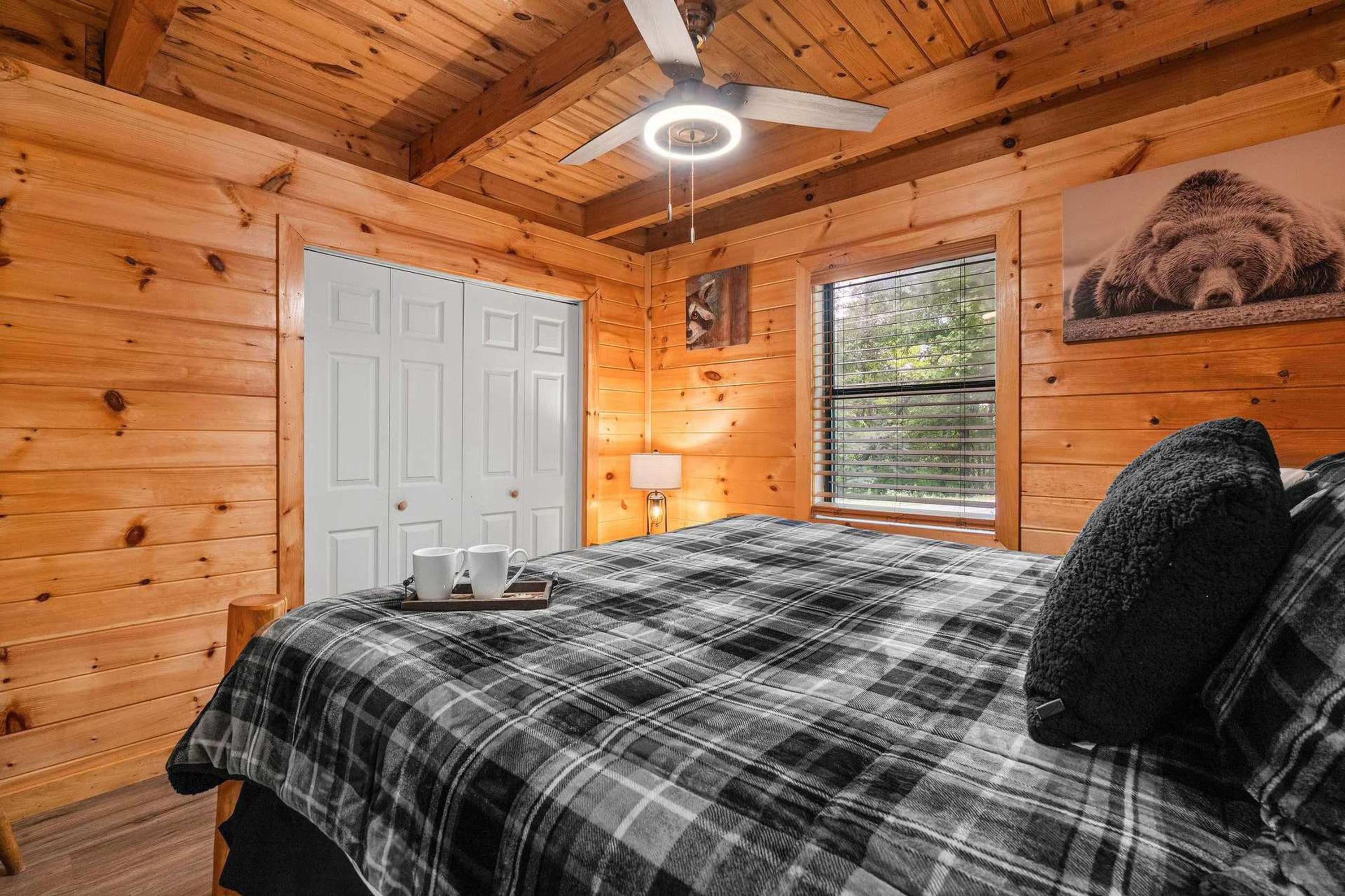 A wooden cabin bedroom with a plaid bedspread, a ceiling fan, a window with blinds, and a bear picture on the wall.