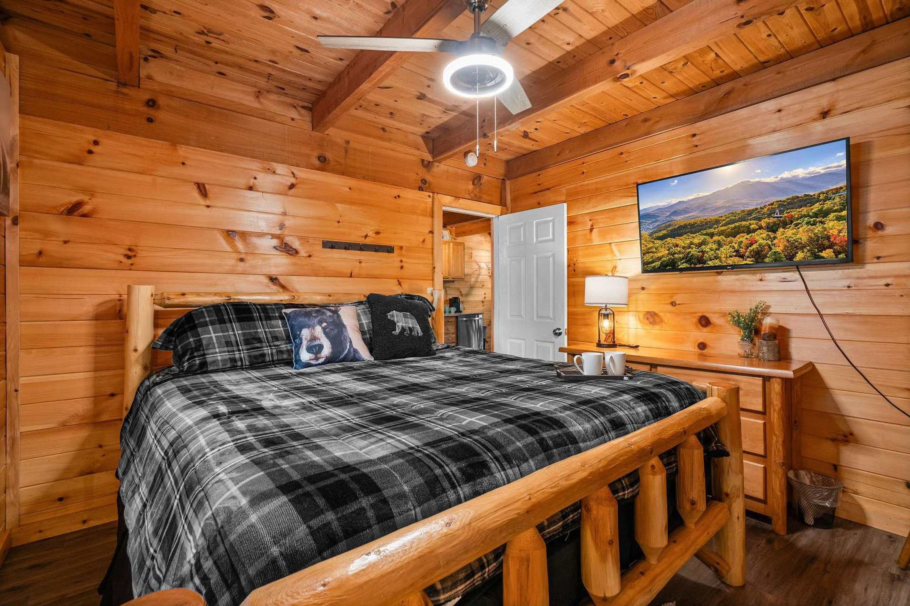 A wooden cabin bedroom with a bed, a ceiling fan, a mounted TV, and a door leading to another room.