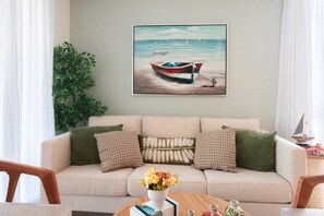 Ein Wohnzimmer mit einem beigen Sofa, grünen und beigen Kissen, einem Gemälde eines Bootes auf dem Wasser, einer Topfpflanze und einem kleinen Tisch mit einer Blumenvase.