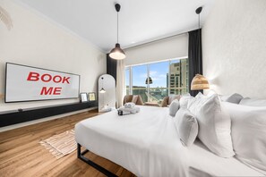 Ein modernes Hotelzimmer mit einem großen Bett, einem 'BOOK ME'-Schild, Blick auf die Stadt und einem großen Fenster.