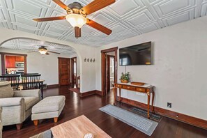 A spacious living room with a ceiling fan, a flat-screen TV mounted on the wall, and a dining area with wooden furniture. - Sleeps 7, 2BR-2BA | 5 mins walk to ISU (Ames)