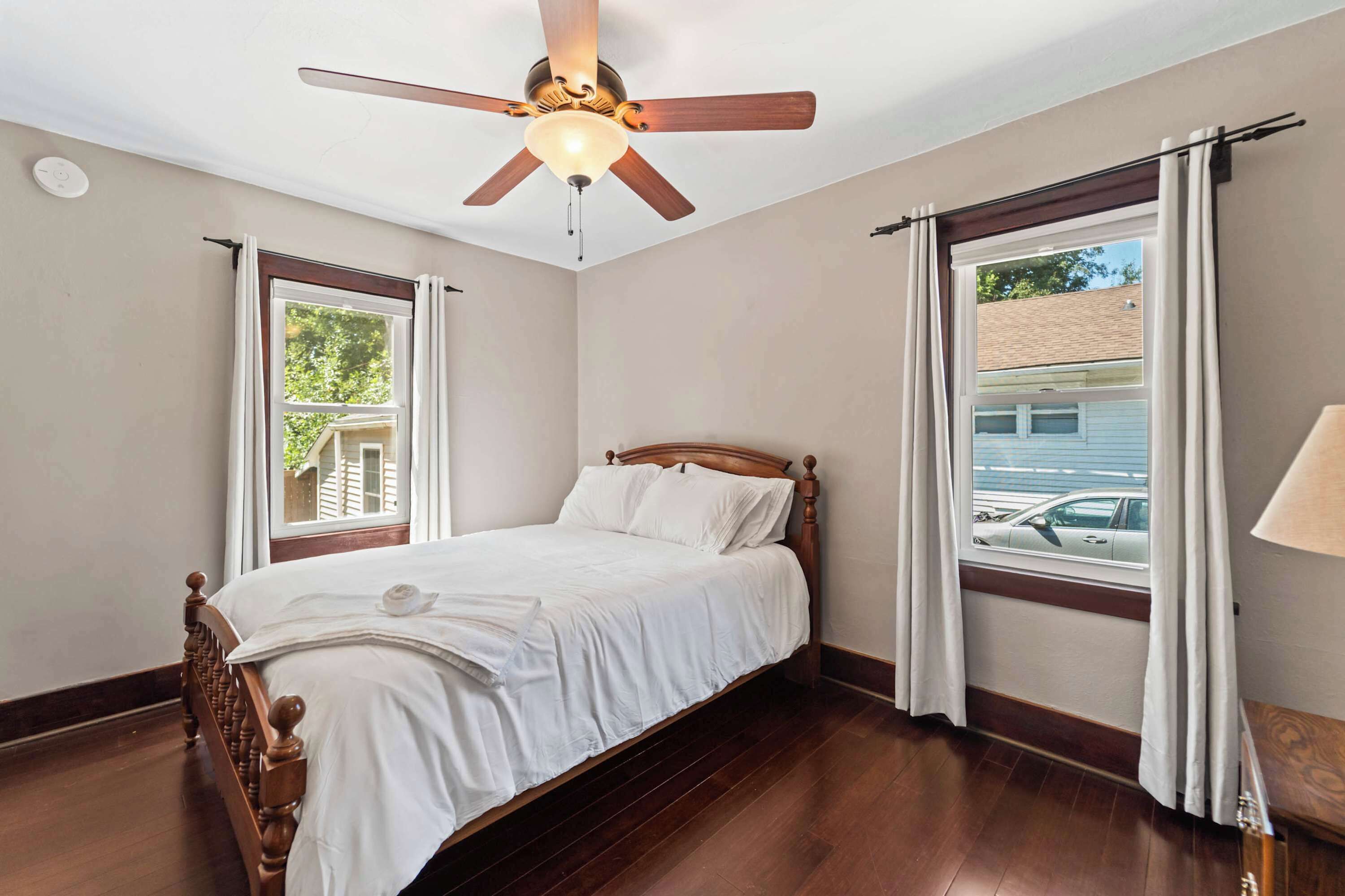 Une chambre à coucher avec un lit en bois, un ventilateur de plafond et deux fenêtres avec des rideaux.