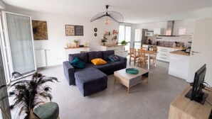 A modern living room with a sofa, coffee table, and dining area. There is a kitchen in the background with white cabinets and a microwave. - L Azulejo - Work From Home Ideal - Mobility Lease Rental - 4pax (Toulouse)