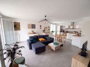 A modern living room with a sofa, coffee table, and dining area. There is a kitchen in the background with white cabinets and a microwave. - L Azulejo - Work From Home Ideal - Mobility Lease Rental - 4pax (Toulouse)