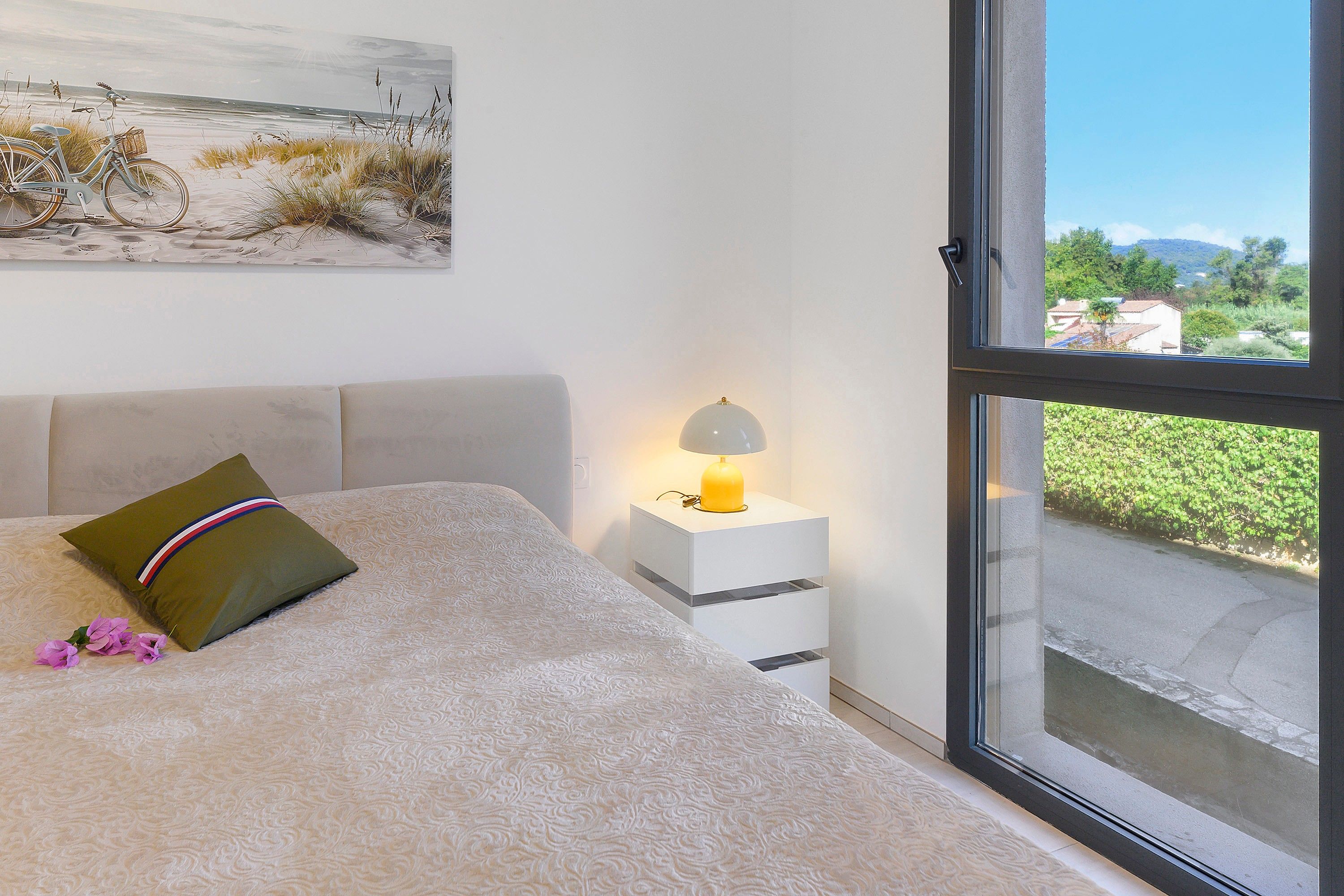 A bedroom with a bed, a nightstand with a lamp, a window with a view of greenery, and a painting on the wall.