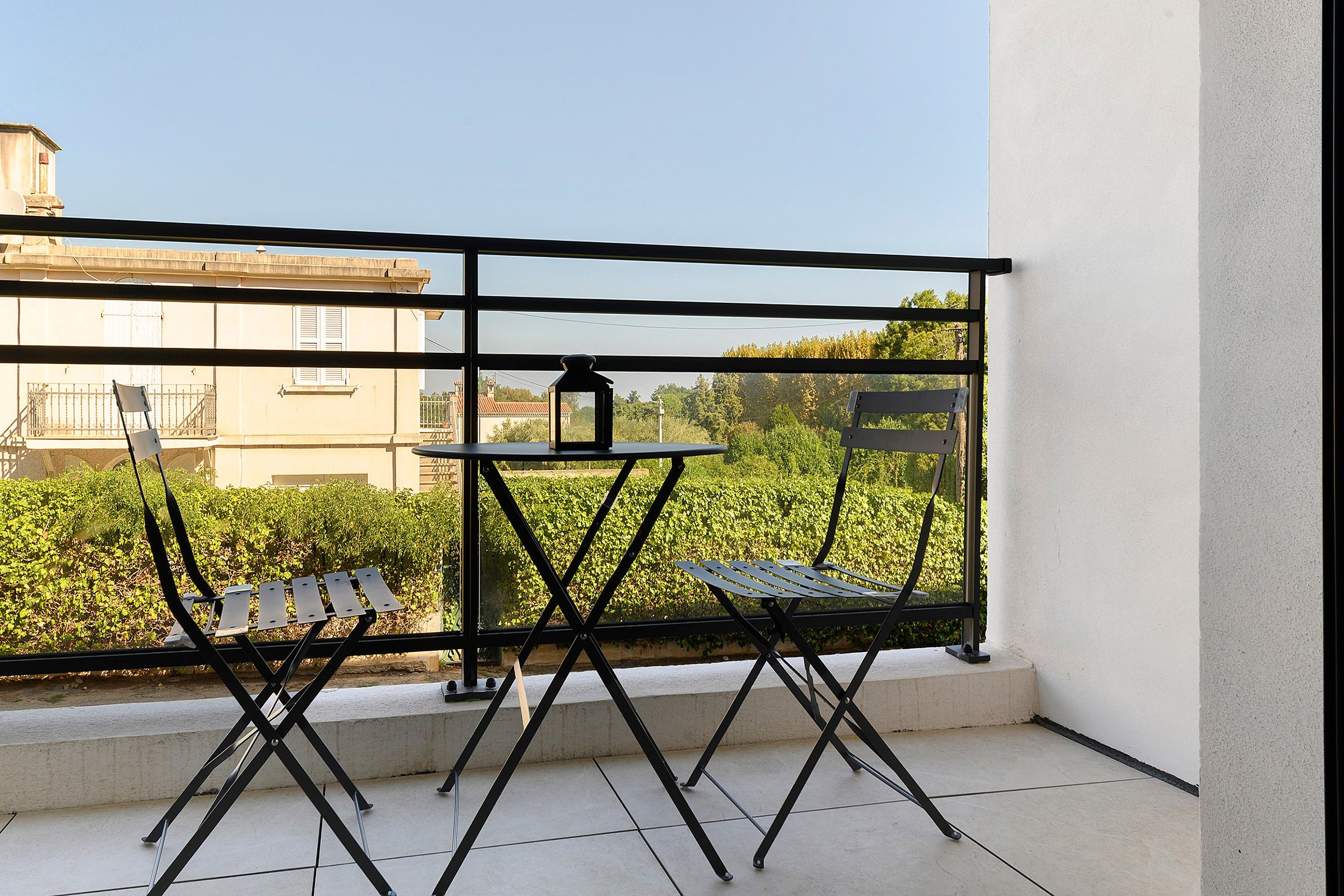 Ein Balkon mit Tisch und zwei Stühlen, einer Laterne und Blick auf Gebäude und Grünflächen.