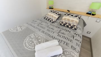 A neatly made bed with a geometric patterned duvet and pillows, a bedside table with a lamp, and a wall-mounted shelf with a phone and decorative items.