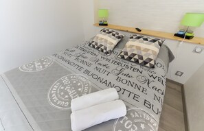 A neatly made bed with a geometric patterned duvet and pillows, a bedside table with a lamp, and a wall-mounted shelf with a phone and decorative items. - Room 'Chambre Brésil' with Shared Garden and Wi-Fi (Rizaucourt-Buchey)
