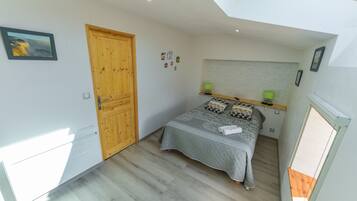 A bedroom with a wooden door, a bed with a quilted cover, a headboard with a shelf, and a small window.