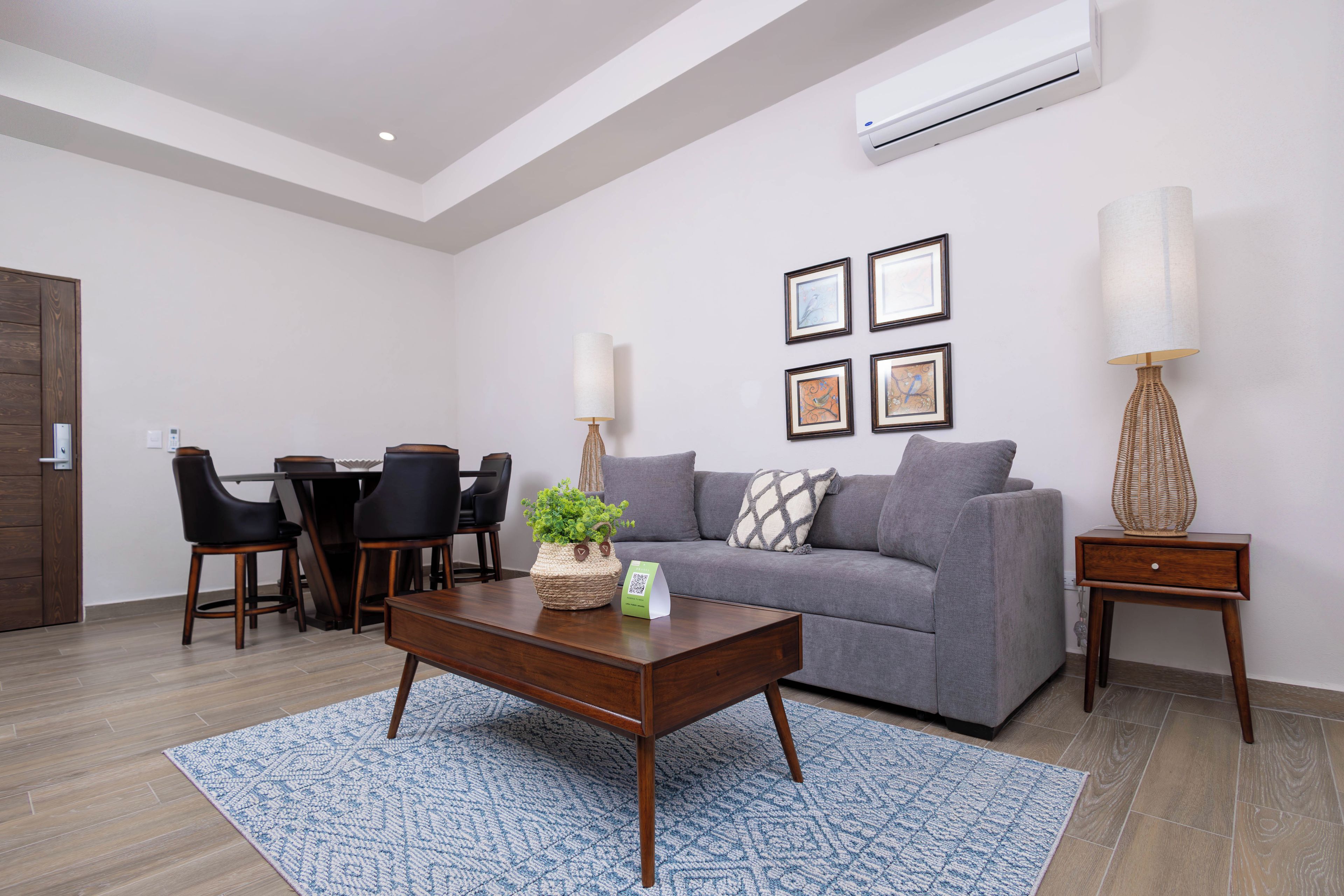 Una sala de estar moderna con un sofá gris, una mesa de centro, un área de comedor con sillas y un aire acondicionado instalado en la pared.