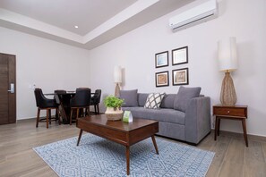 A modern living room with a grey sofa, a coffee table, a dining area with chairs, and a wall-mounted air conditioner.
