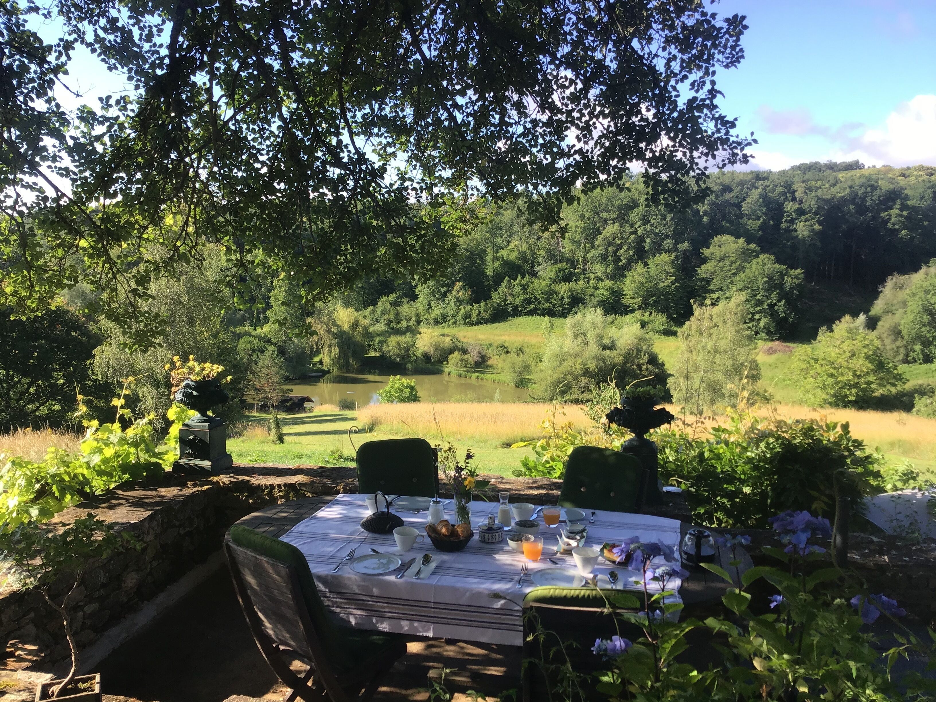 Un área de comedor al aire libre con una mesa dispuesta para dos personas, rodeada de vegetación y vista a un lago.