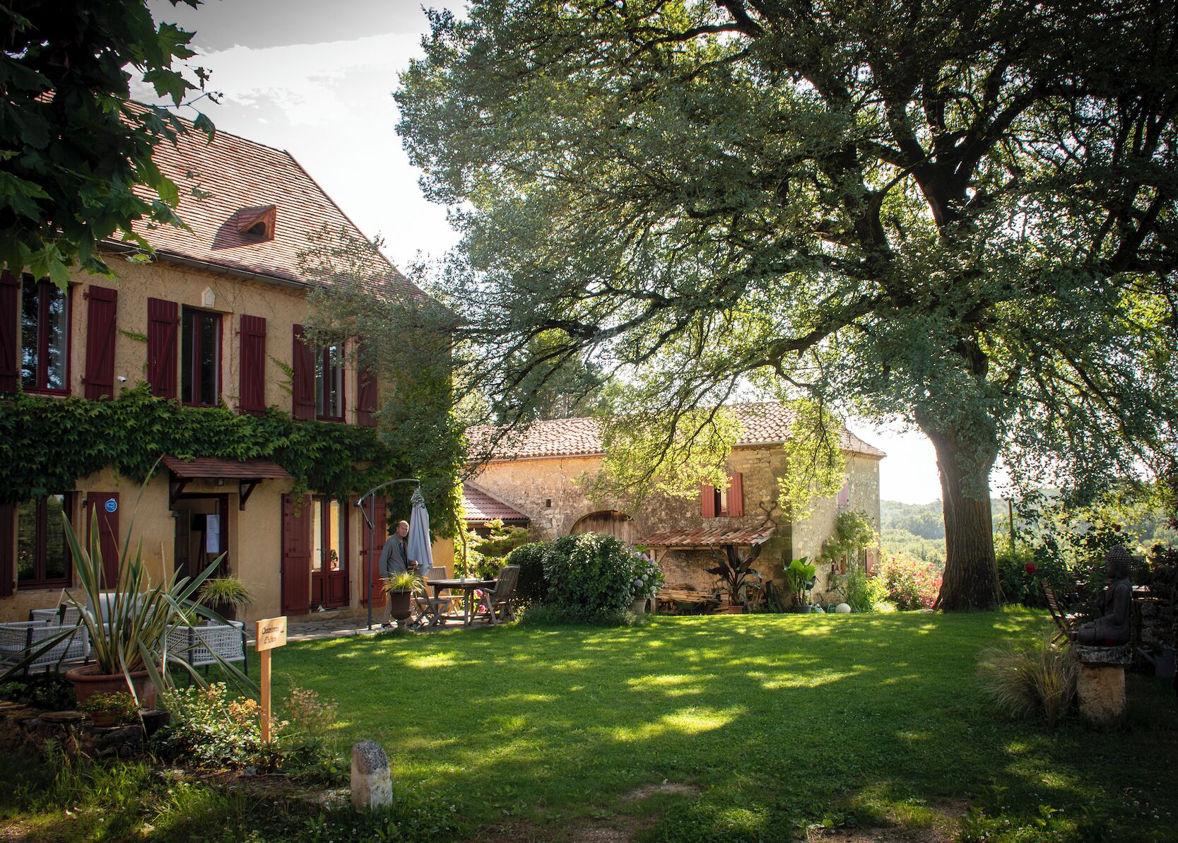 Un bâtiment traditionnel en pierre, doté d’un jardin, d’un banc et d’un arbre qui offre de l’ombre.