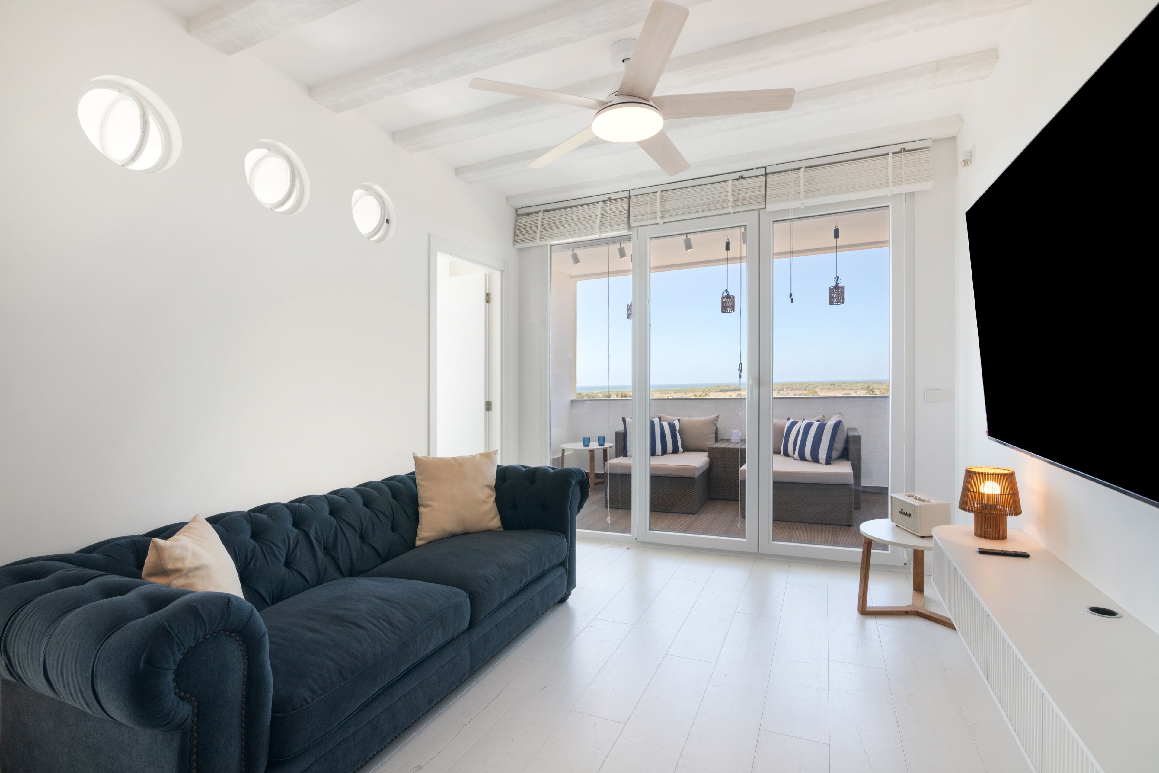 Un salon moderne avec un canapé bleu foncé, un ventilateur de plafond et une grande fenêtre donnant sur une plage.