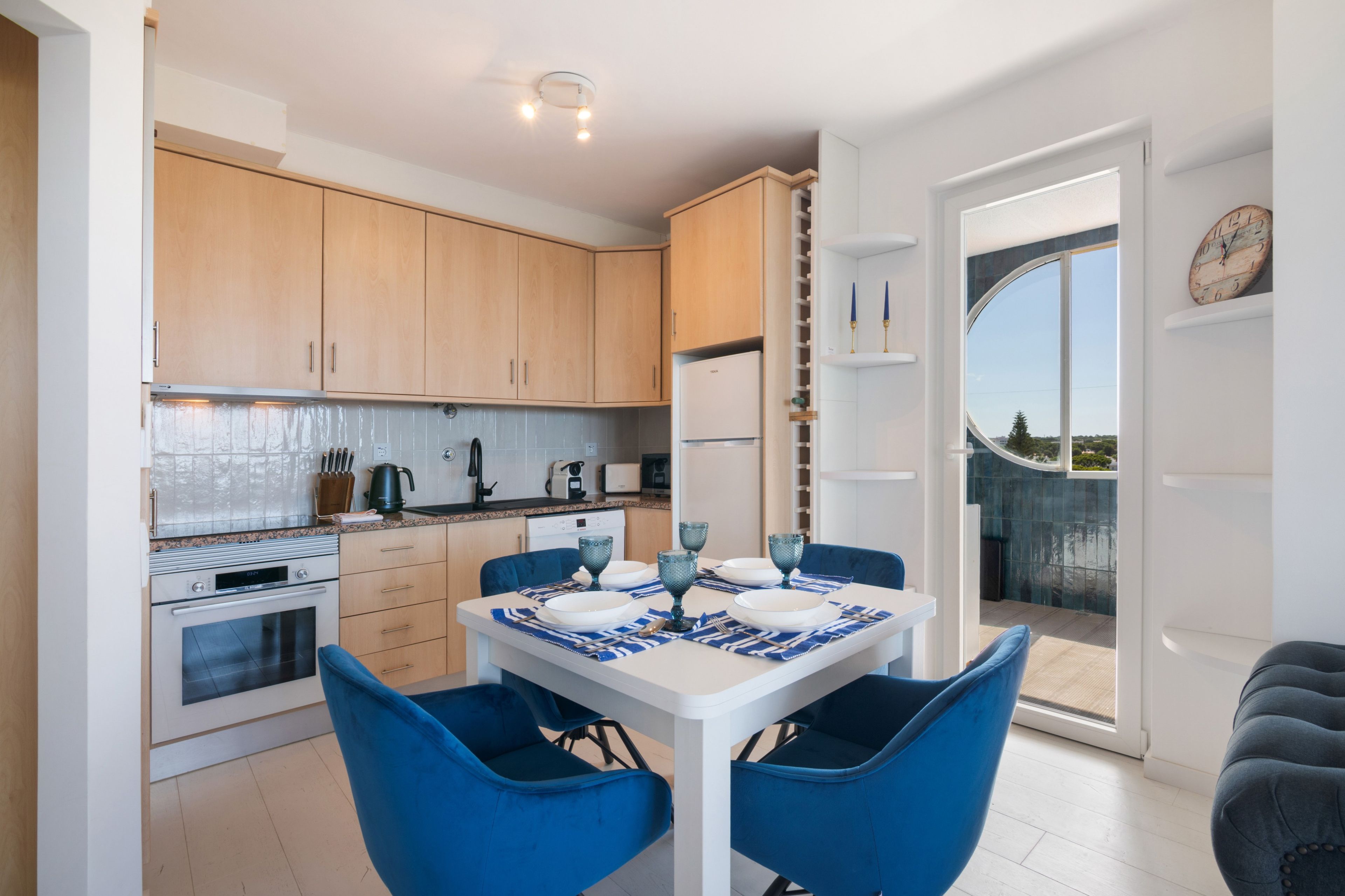 Une cuisine moderne avec des meubles blancs, une table à manger entourée de chaises bleues et une vue sur l’extérieur grâce à une porte vitrée.