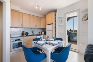 A modern kitchen with white cabinets, a dining table set with blue chairs, and a view of the outdoors through a glass door.