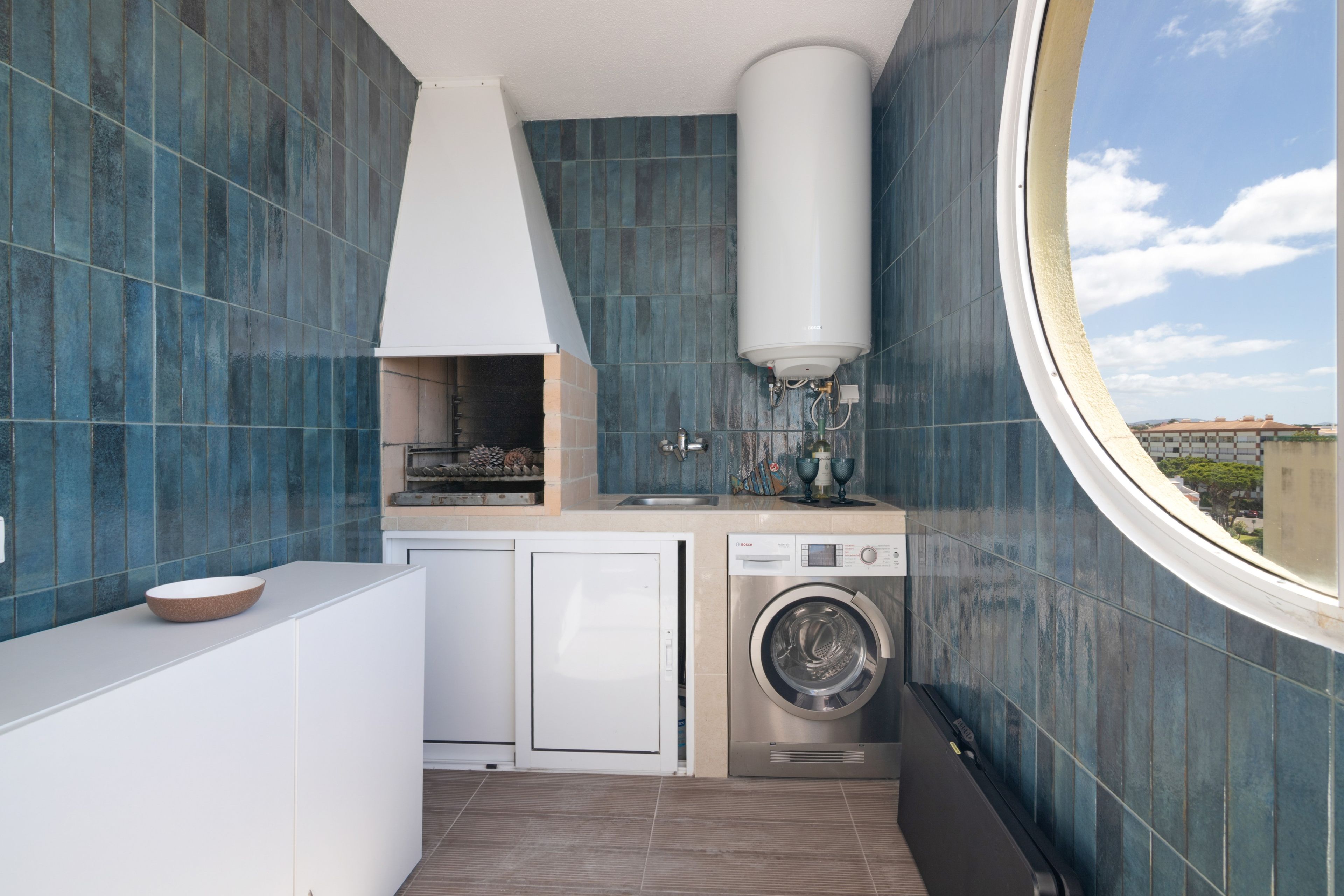 A compact kitchen with a built-in oven, a washing machine, and a window overlooking a cityscape.