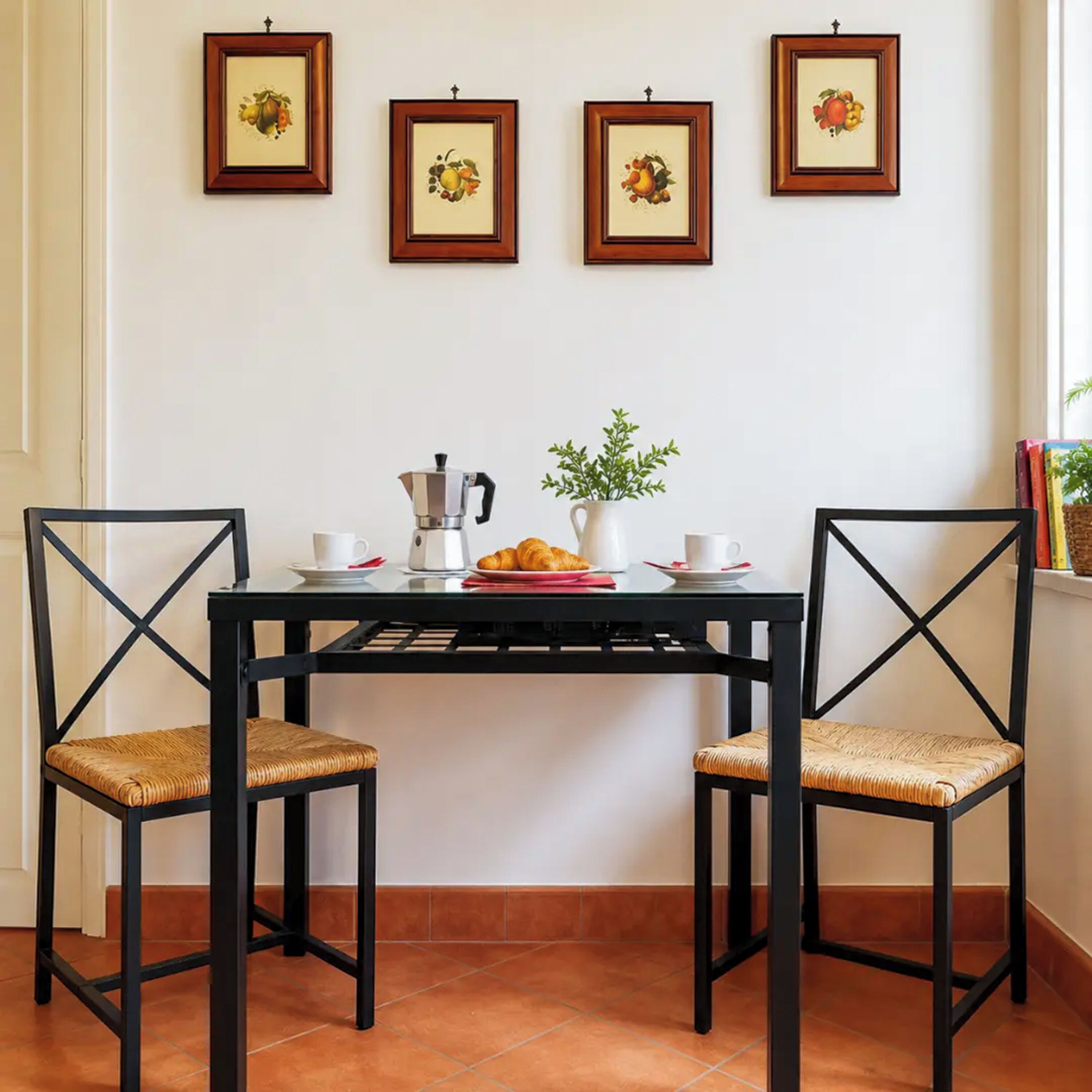 A dining area with a black table, two chairs, a coffee maker, a vase with green plants, and four framed pictures on the wall.