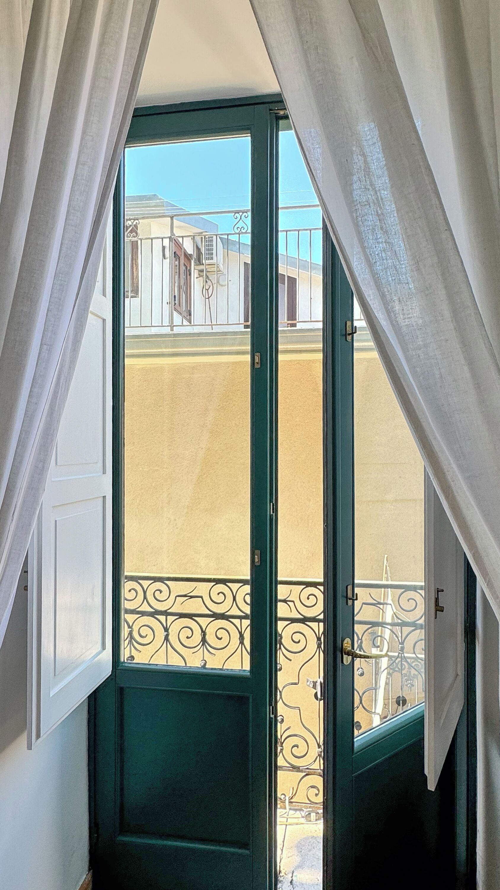 Vista a través de una puerta con marco verde y cortinas blancas, que da a un balcón con barandilla de metal.