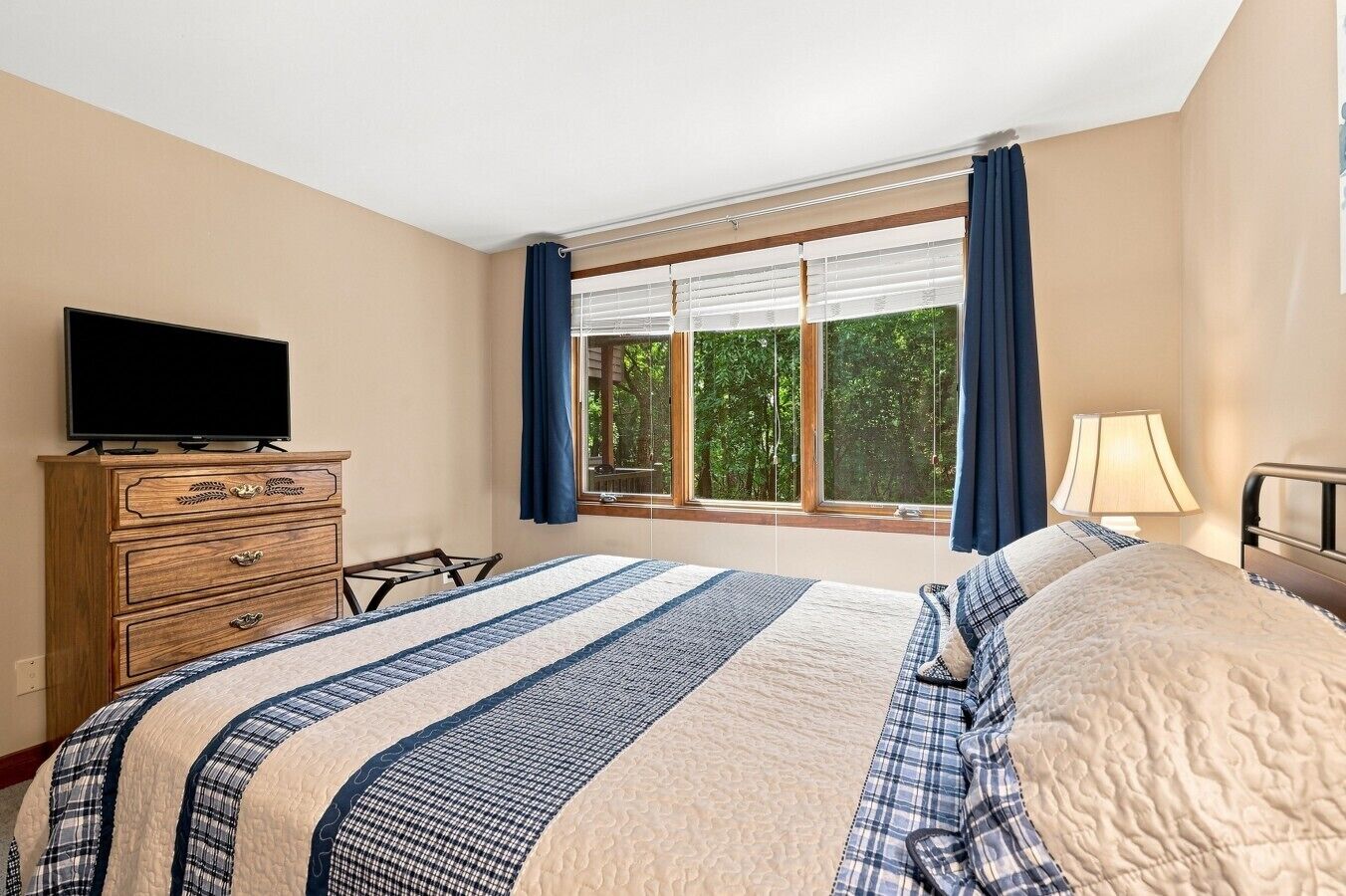 Une chambre à coucher avec un lit, une commode, un téléviseur, une fenêtre avec des rideaux et une lampe.