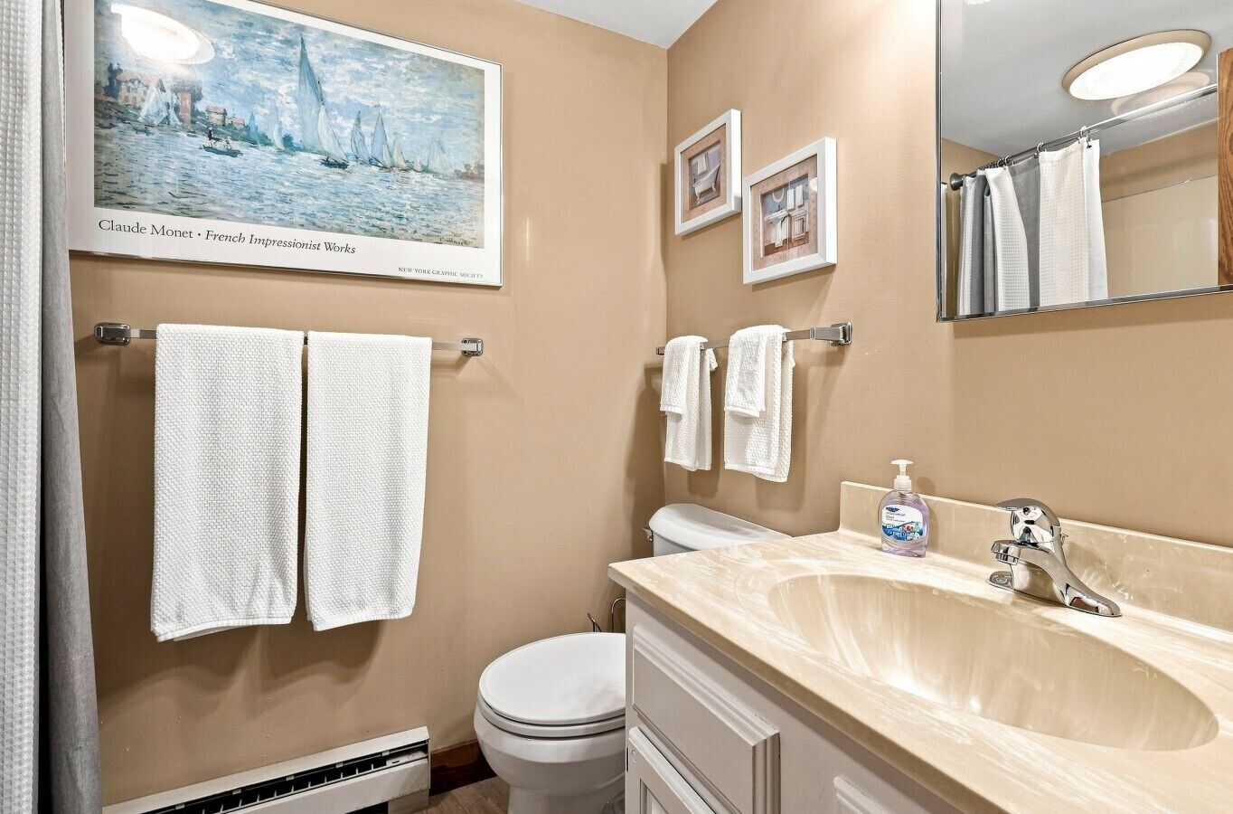 A bathroom with a beige wall, a white toilet, a sink with a mirror, and two framed pictures.