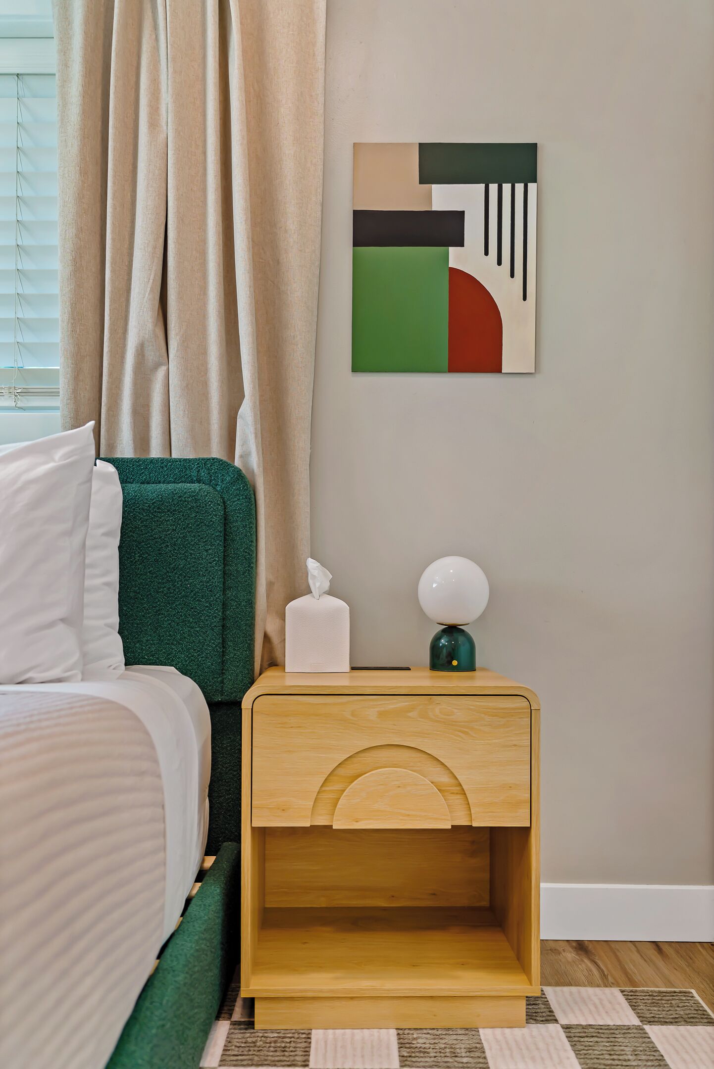 Ein Schlafzimmer mit einem Bett, einem Nachttisch mit Taschentuchbox und Lampe, einer Wand mit einem geometrischen Kunstwerk und einem Fenster mit Vorhängen.
