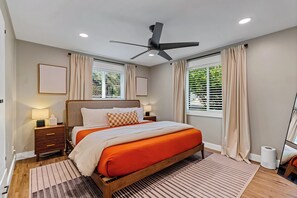 Un dormitorio moderno con cama, mesitas de noche, ventilador de techo y ventanas con cortinas.