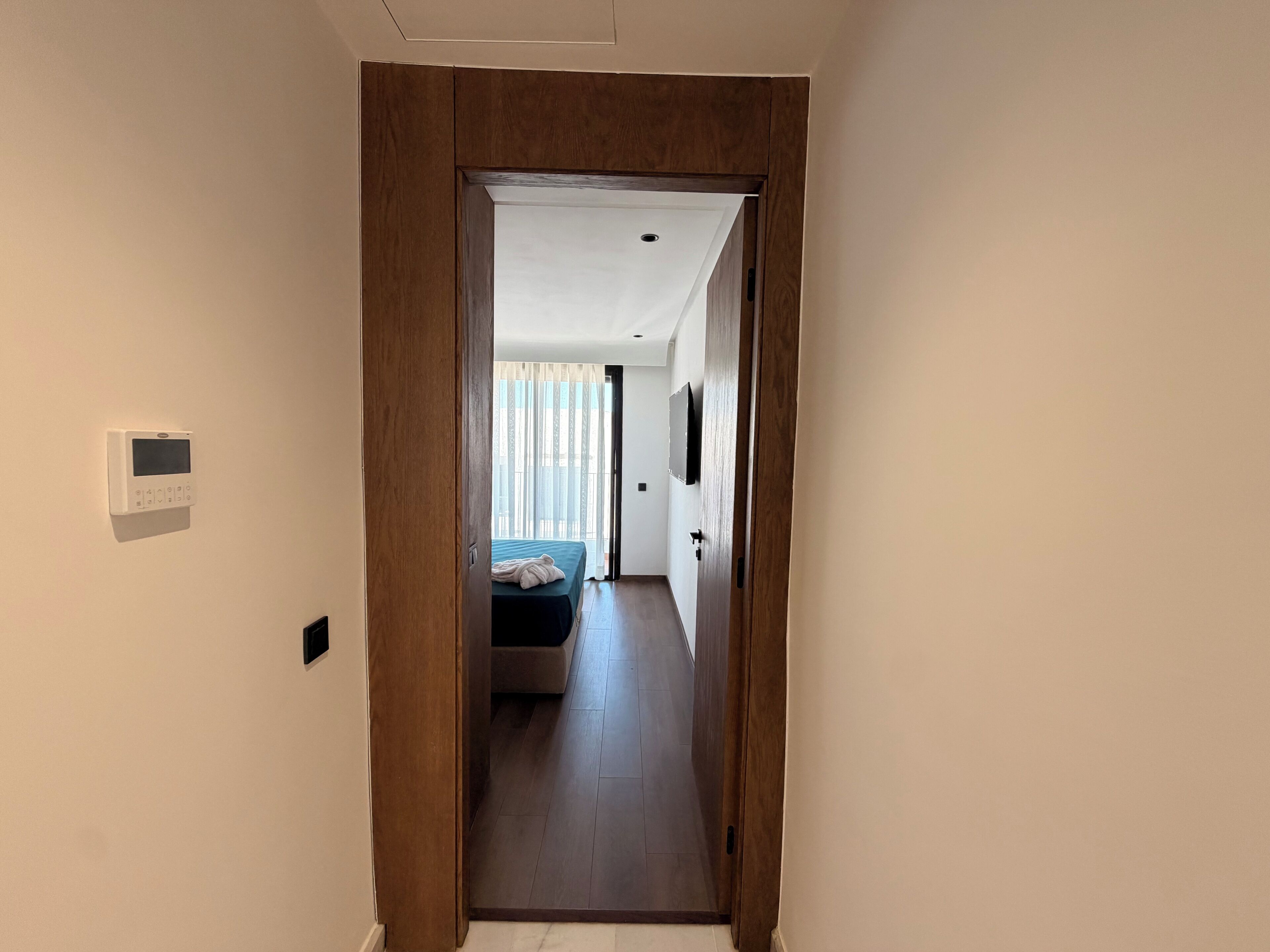 Un pasillo que conduce a un dormitorio con una cama, un termostato en la pared y una puerta con marco de madera.