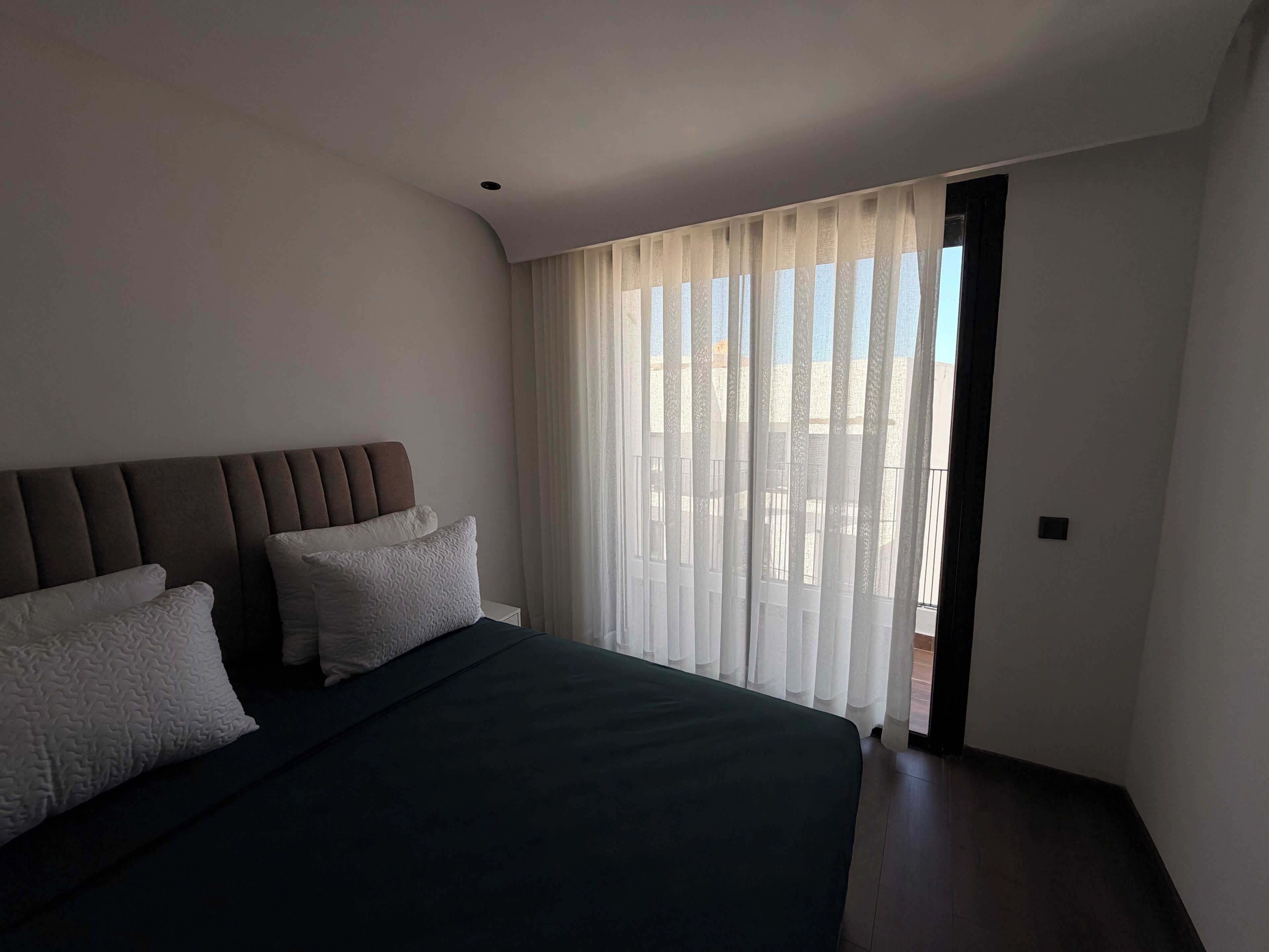 Un dormitorio con cama, cabecera y ventana con cortinas translúcidas.