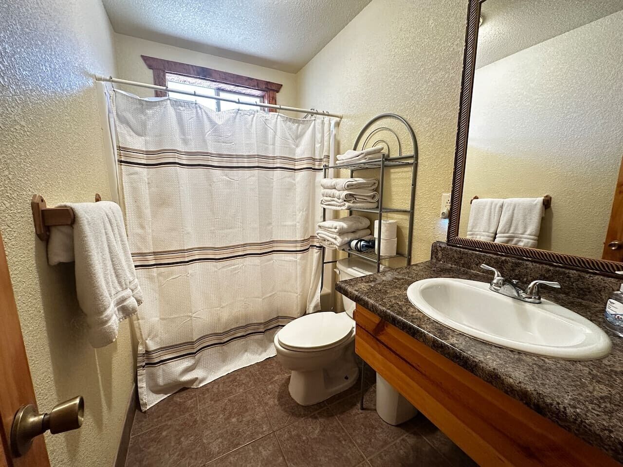 Un baño con cortina de ducha, inodoro, lavamanos con espejo y toallas en un perchero.