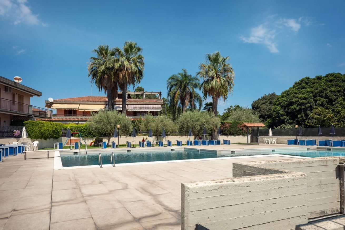 Zona de piscina amb gandules i ombrelles, envoltada de palmeres i edificis.