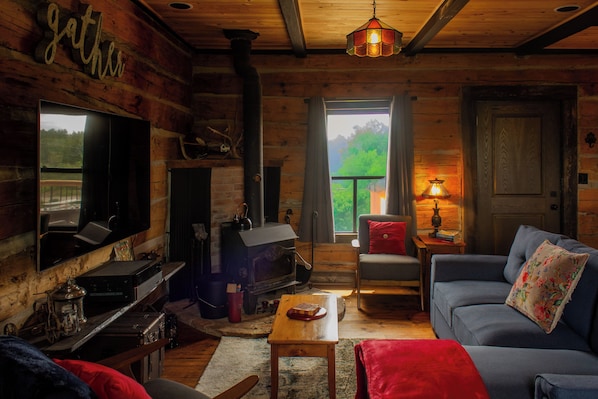 A log cabin interior with a fireplace, sofa, television, and a view of the outdoors. - The Lodge | Historic Log Cabin + Hot Tub + Beach Access (Tweed)
