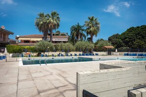 Zona de piscina amb gandules i ombrel·les, envoltada de palmeres i un edifici amb balcons.