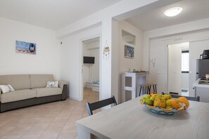 A modern living room with a sofa, a dining table with fruit, and a kitchen area. - GuestHost - BaiAmare - Apartment with Shared Pool (Capo Mulini)