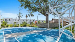 Una cancha de baloncesto con superficie azul, rodeada de árboles y un edificio blanco al fondo.