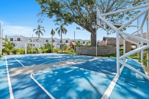 Una cancha de baloncesto con superficie azul, rodeada de árboles y un edificio blanco al fondo.