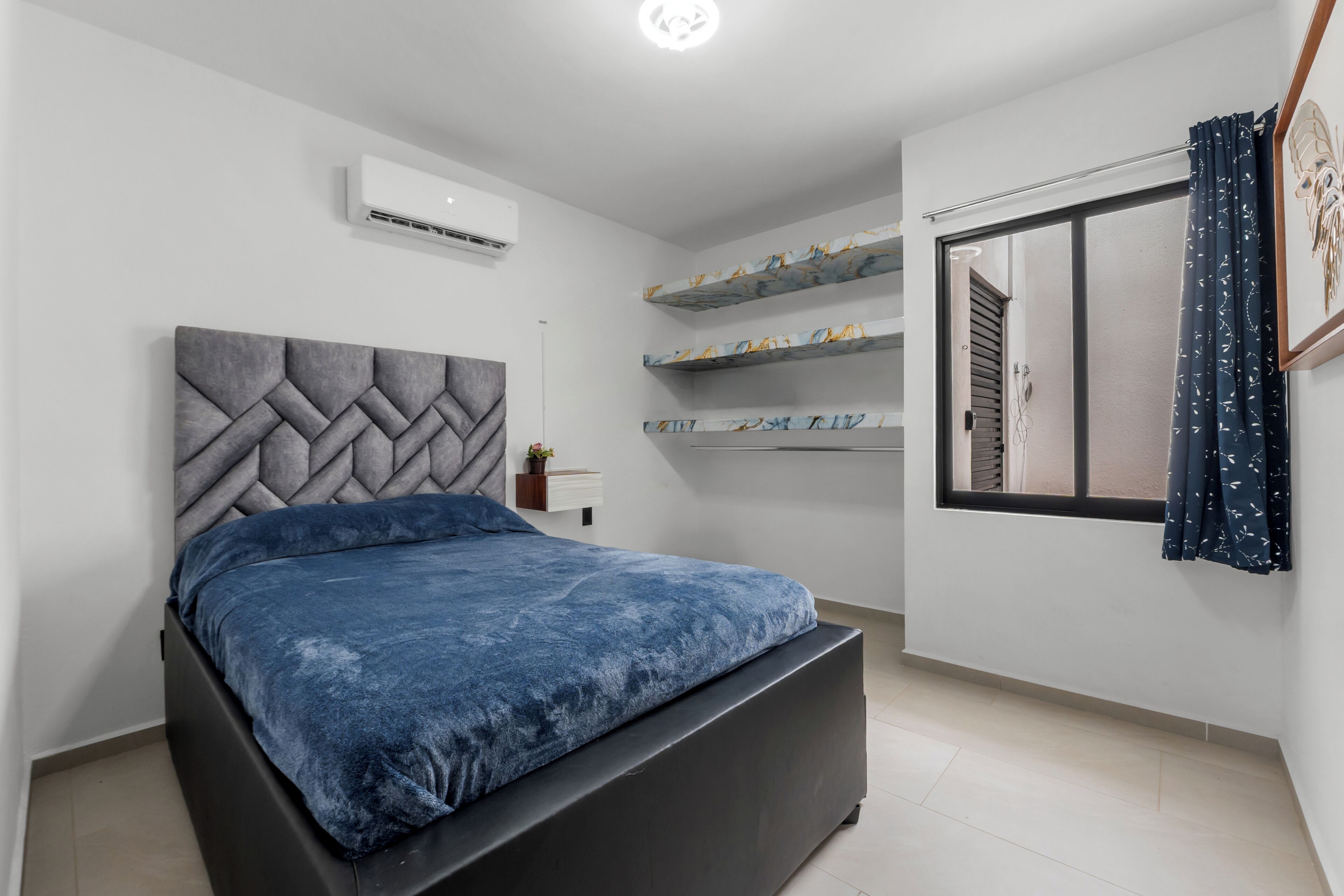 Un dormitorio con cama, aire acondicionado de pared, ventana con cortinas y estanterías empotradas con objetos decorativos.
