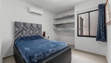 Un dormitorio con cama, aire acondicionado de pared, ventana con cortinas y estanterías empotradas con objetos decorativos.