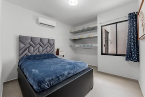 A bedroom with a bed, a wall-mounted air conditioner, a window with curtains, and built-in shelves with decorative items.