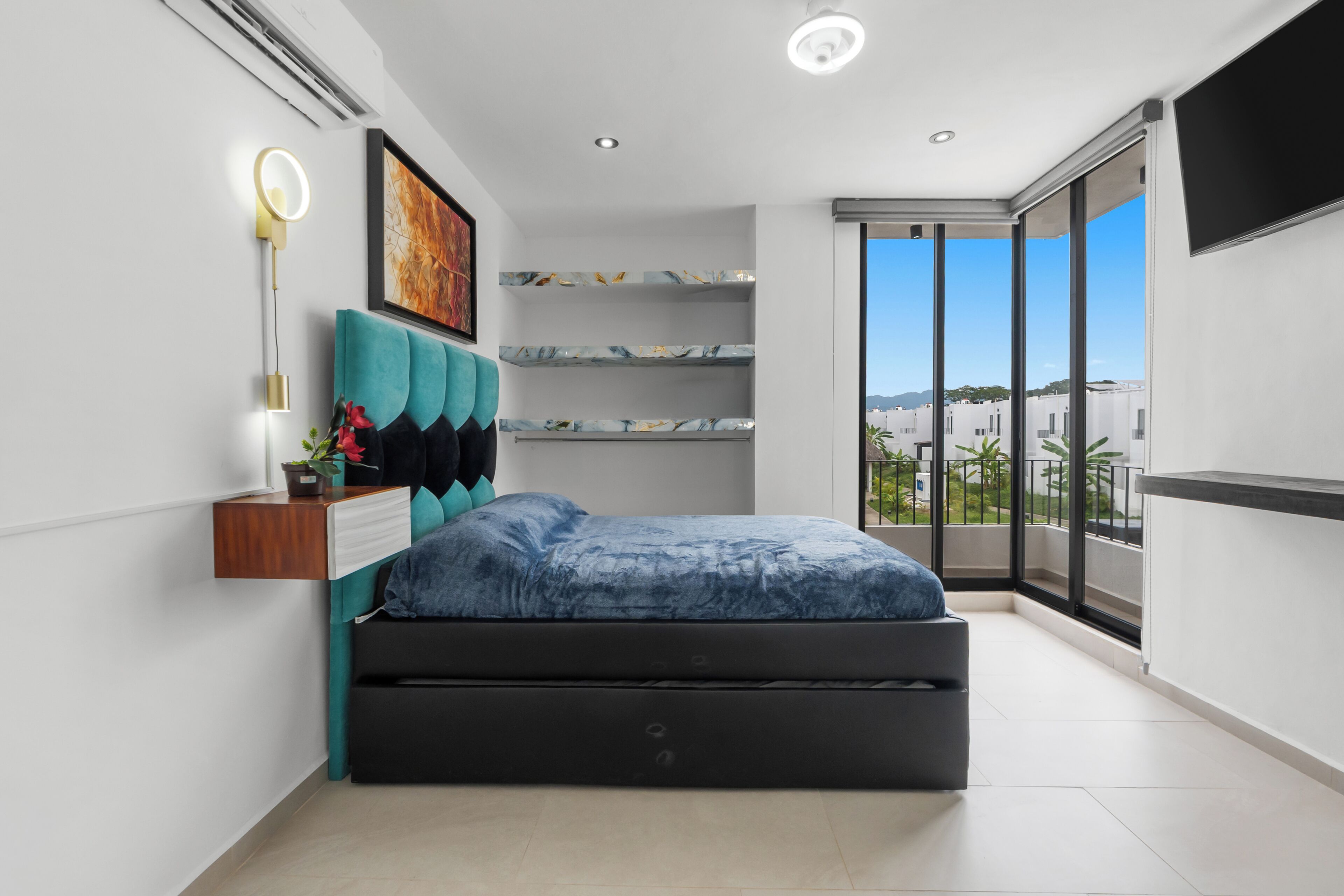 A modern bedroom with a bed, a bedside table, a wall-mounted shelf, and a large window.