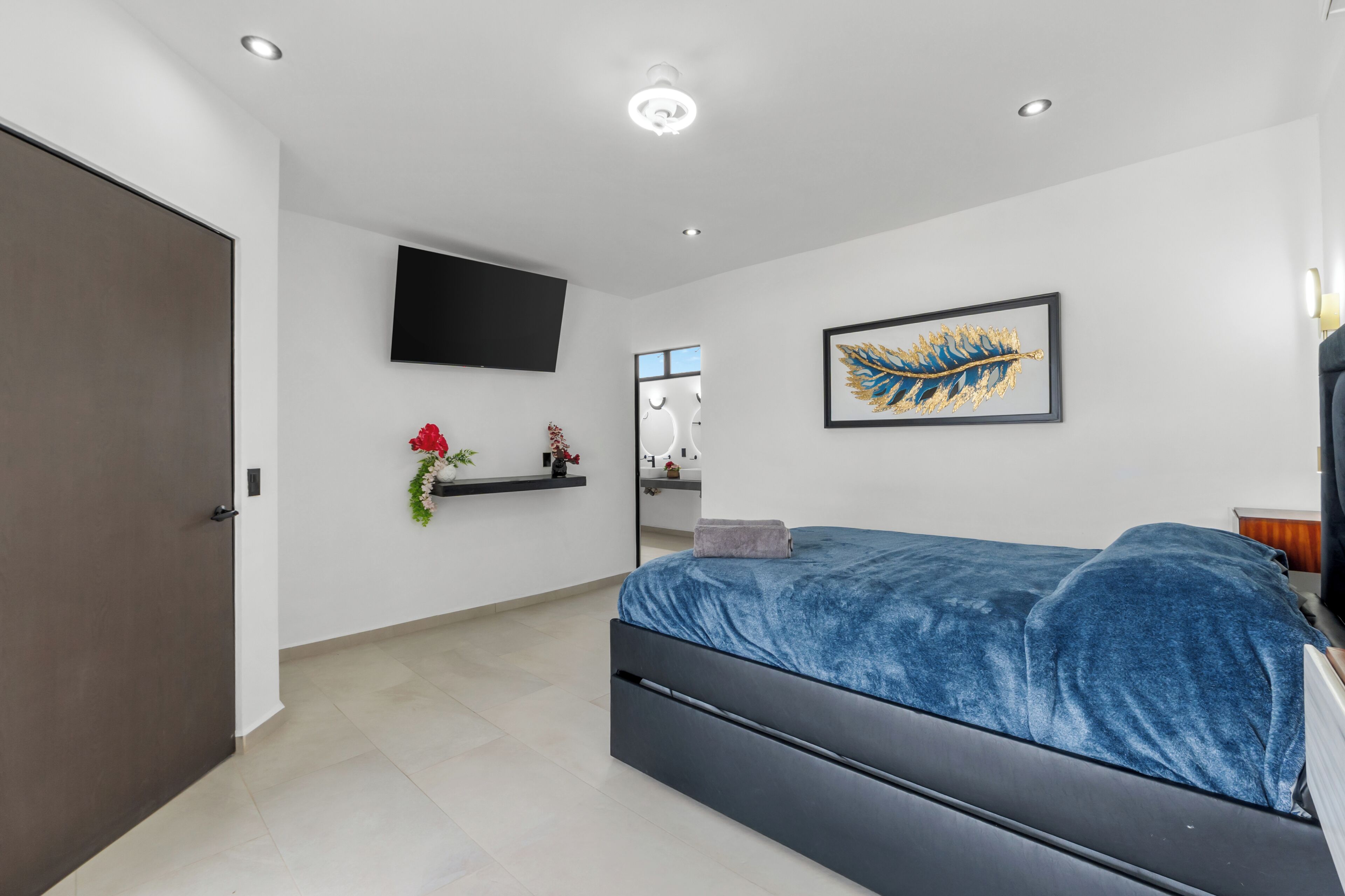 A modern bedroom with a bed, a mounted TV, a wall-mounted artwork, and a bathroom visible through a glass door.