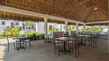 Un área de comedor al aire libre con mesas y sillas, techo de paja y vista a un edificio con ventanas y un automóvil estacionado afuera.