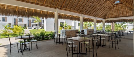 Un área de comedor al aire libre con mesas y sillas, techo de paja y vista a un edificio con ventanas y un automóvil estacionado afuera.