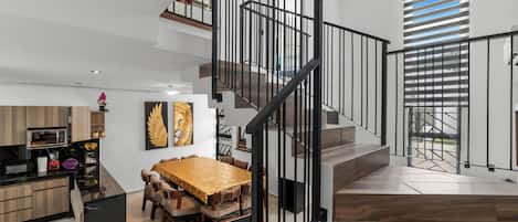 Un interior moderno con una mesa de comedor de madera, una escalera con barandillas negras y una zona de cocina.