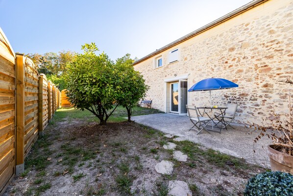 A courtyard with a wooden fence, a small tree, a table with a blue umbrella, and a stone building.