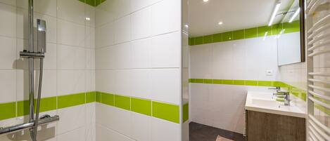 A bathroom with white and green tiles, a shower, a sink with a mirror, and a wooden cabinet.