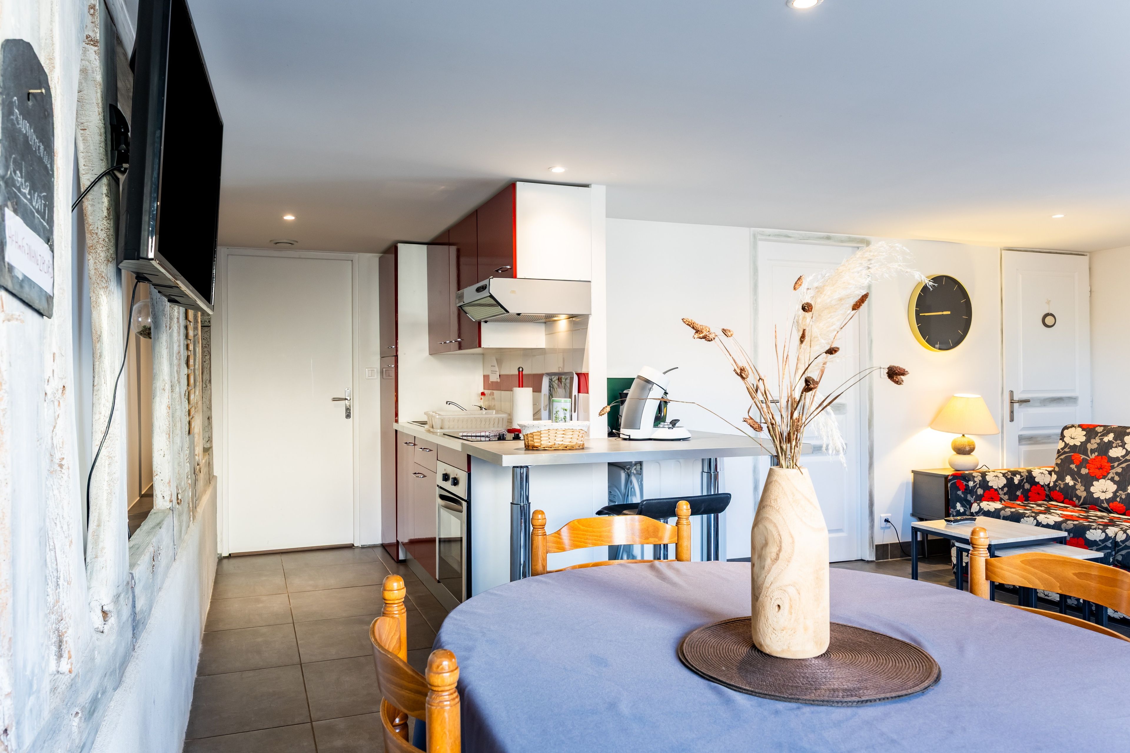 A modern kitchen with a built-in oven, a dining area with a round table, and a living room with a sofa and a wall clock.