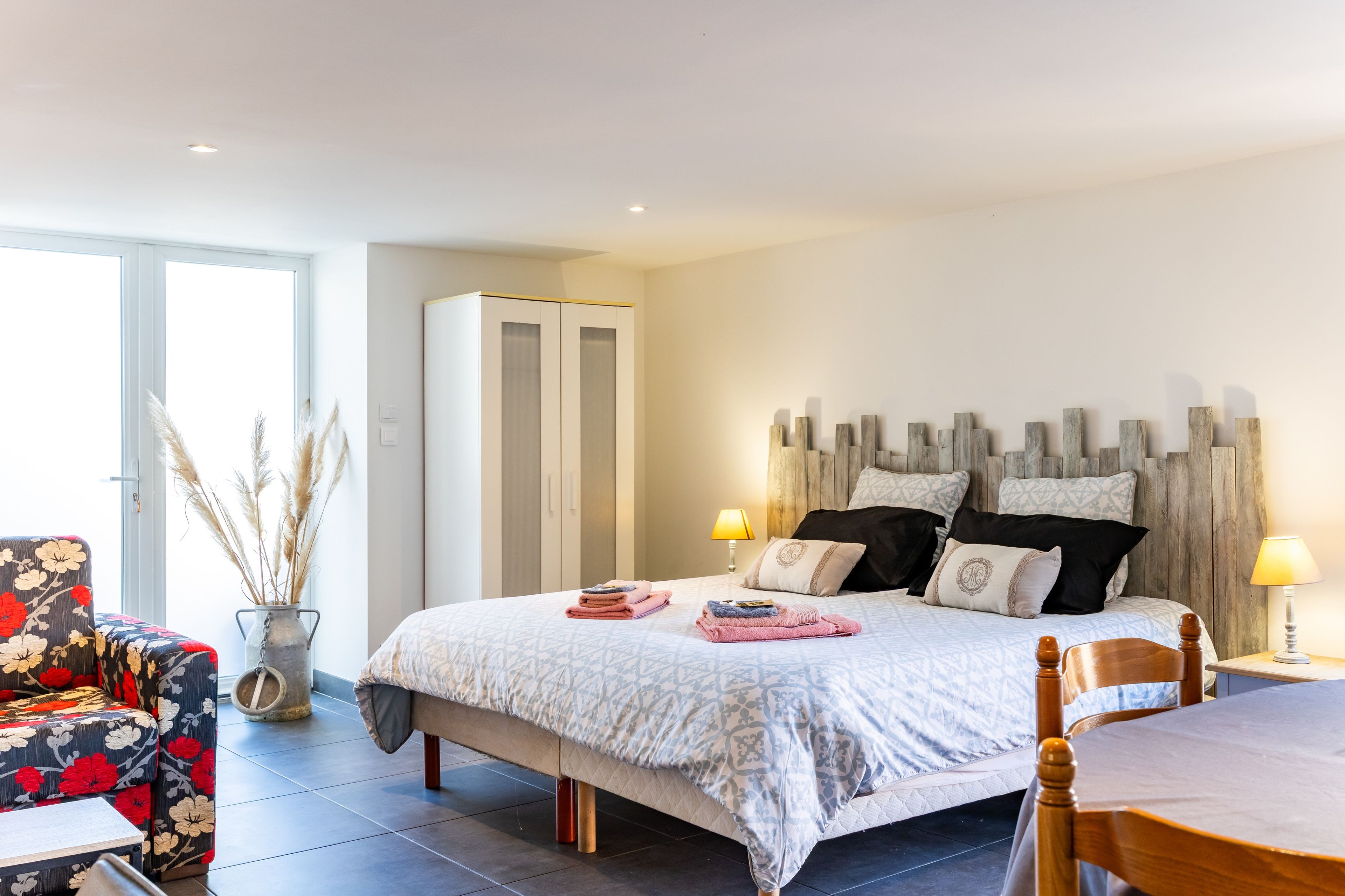 A bedroom with a bed, bedside lamps, a wooden headboard, a chair, a table, and a wardrobe.