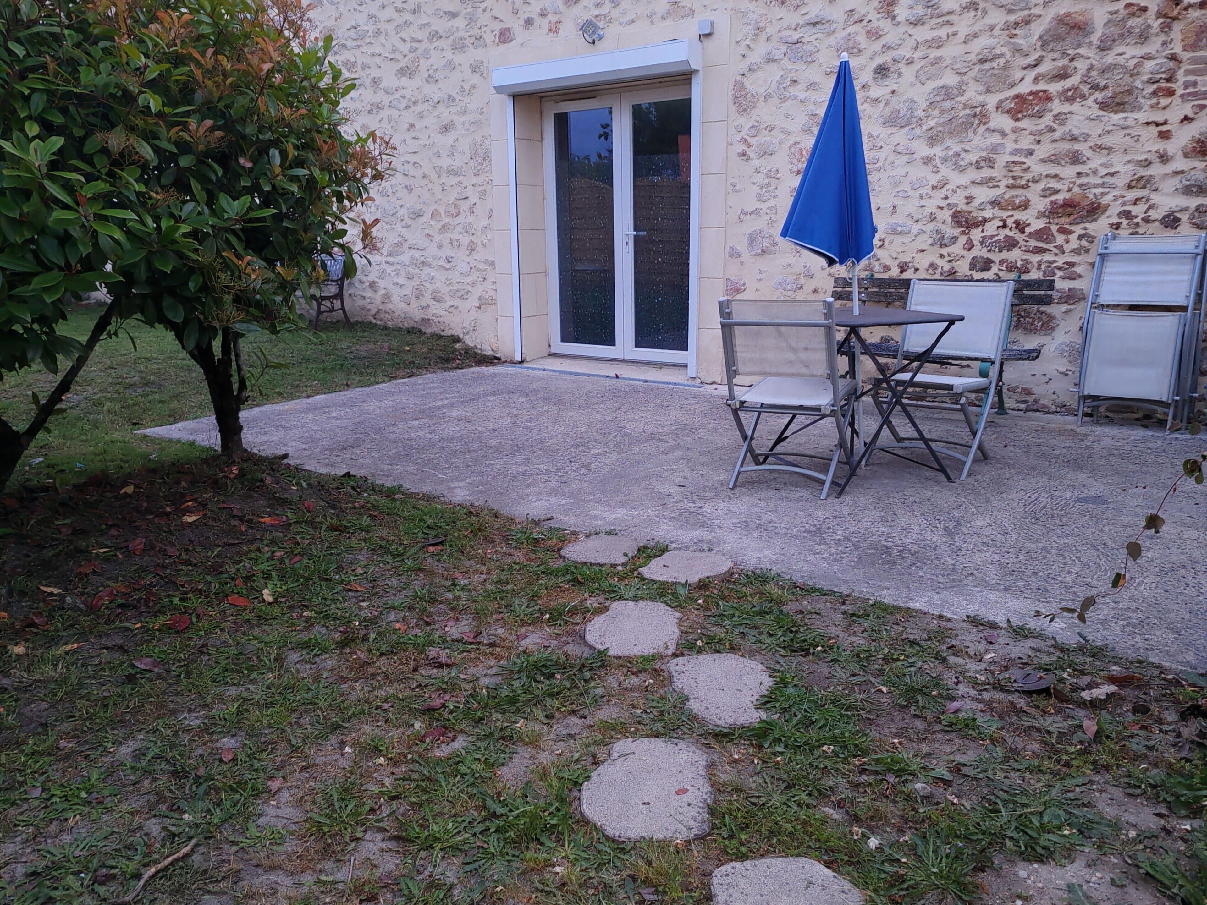 Un espace patio aménagé avec une table et des chaises, un parasol bleu et un chemin de pierre menant à un bâtiment à la façade en pierre.